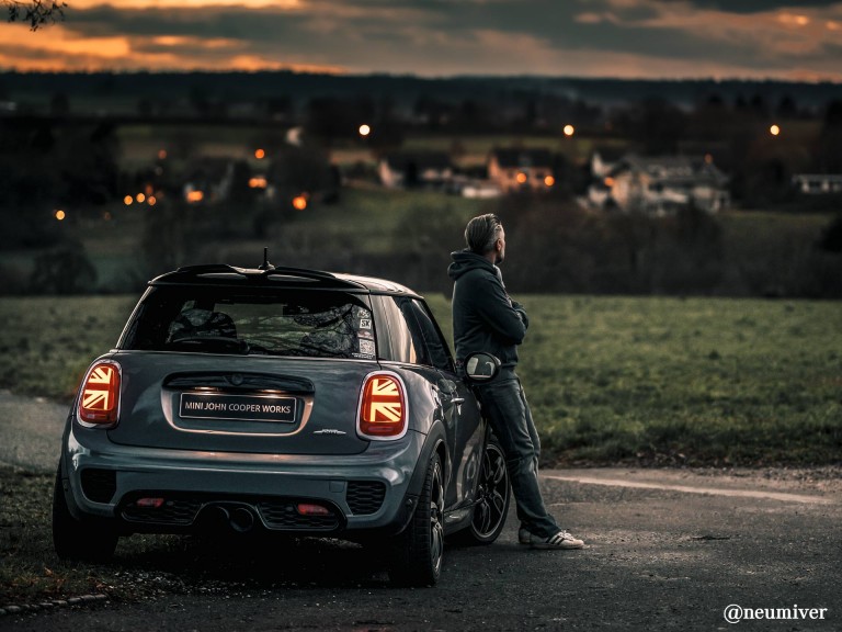 A man standing aside of a MINI COOOPER with city lights in the background - rear view.